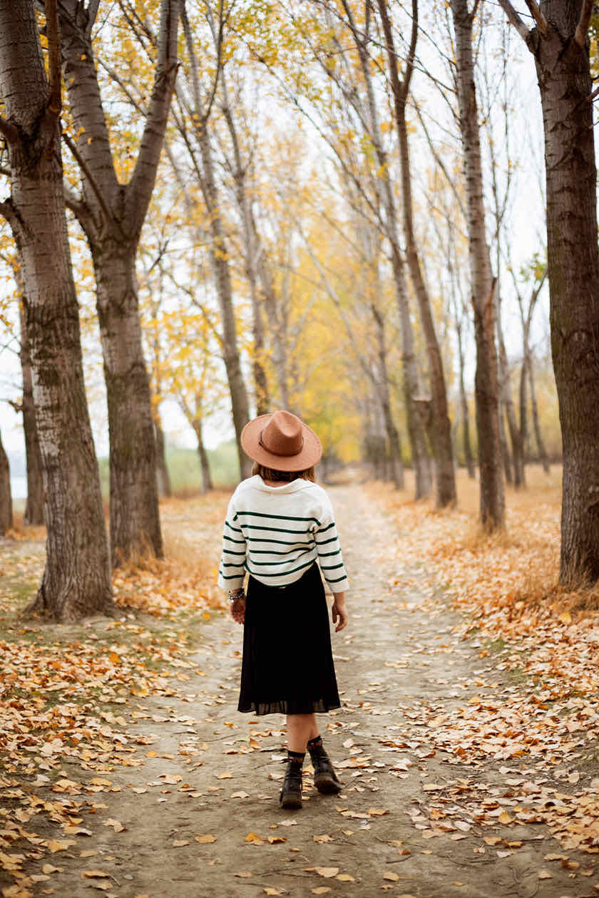Woman in a brown hat and striped sweater walking down a leaf-covered path lined with autumn trees