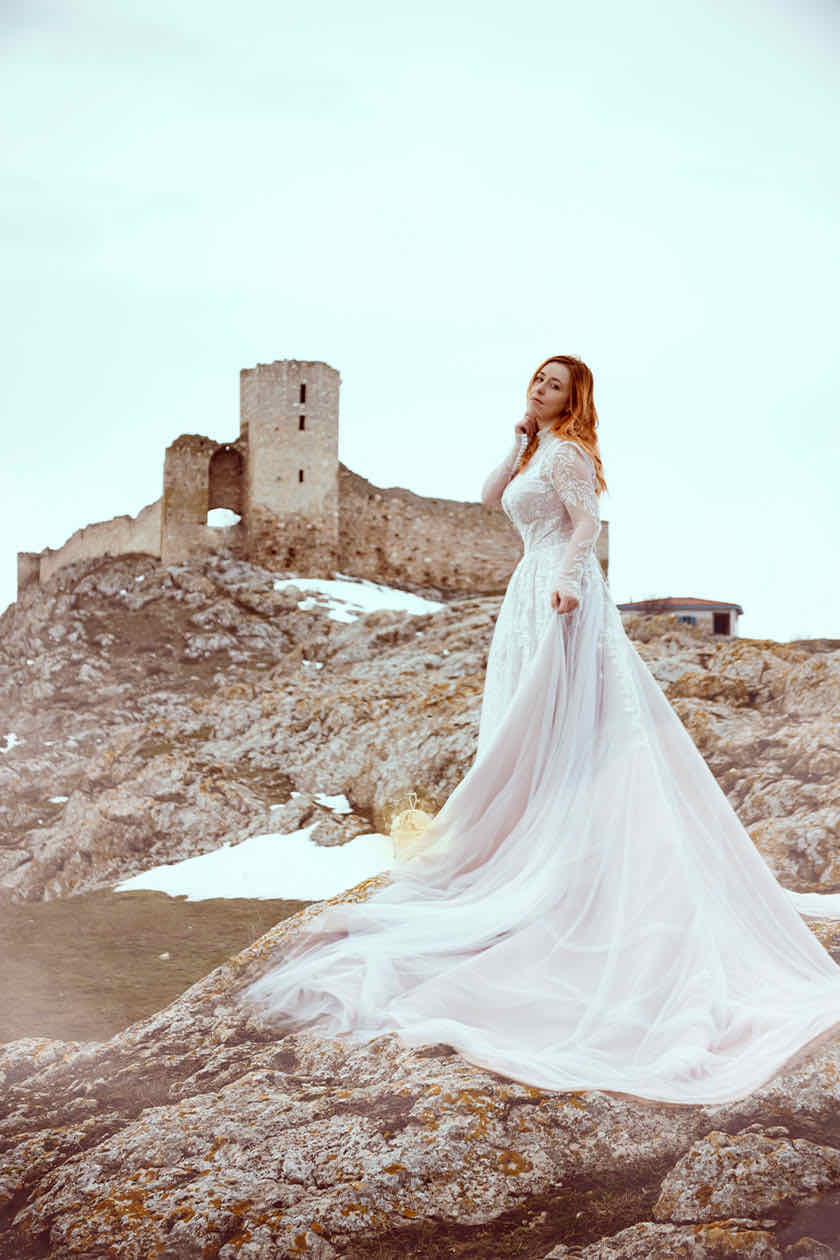 Woman in a white lace wedding gown on a rocky hilltop before medieval castle ruins