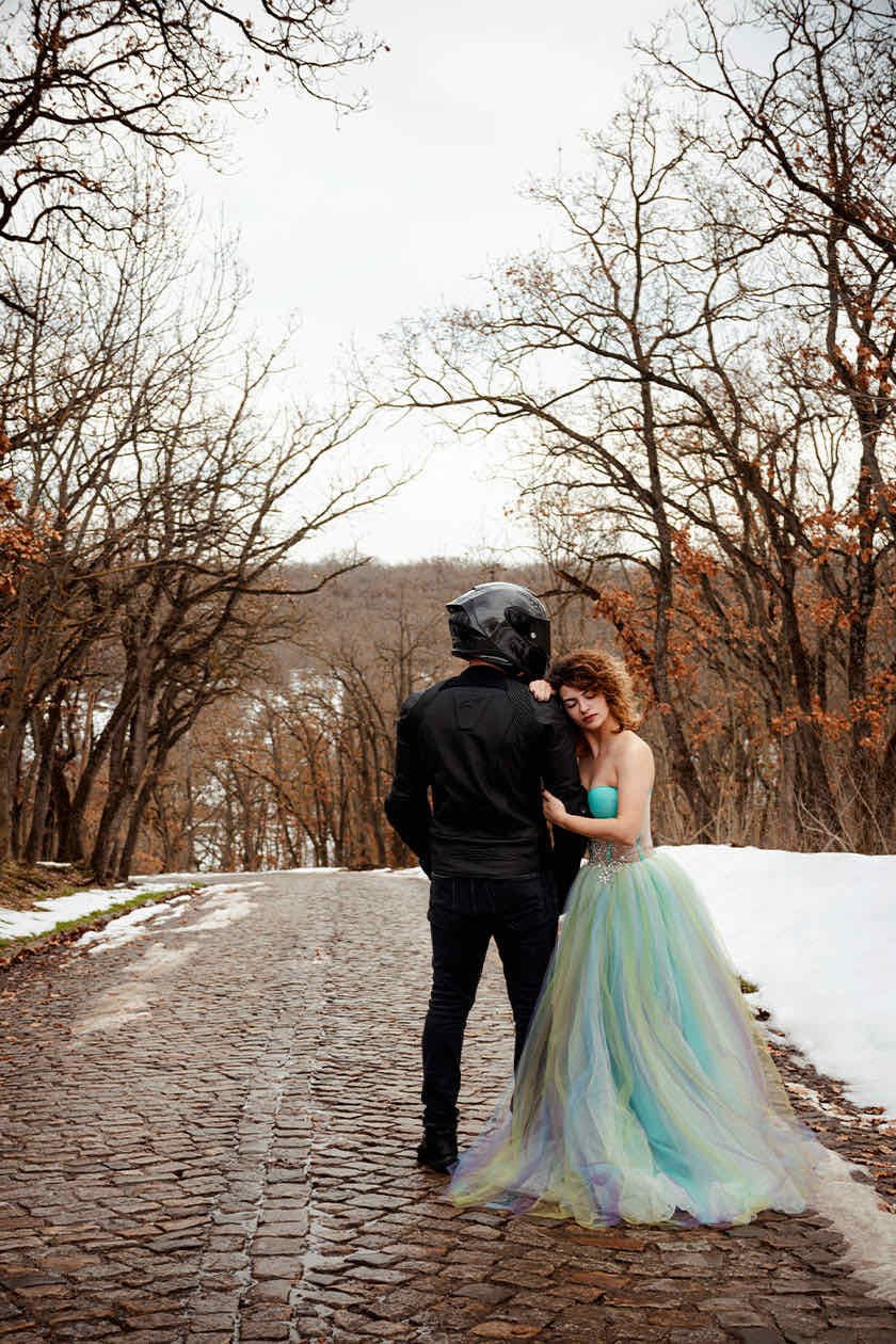 Couple on a cobblestone path in winter, one in a motorcycle jacket and helmet, the other in a pastel tulle gown