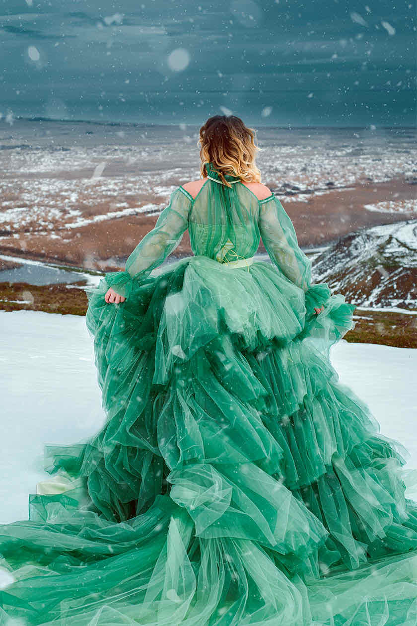 Woman in a voluminous emerald tulle gown looking over a snowy valley during snowfall