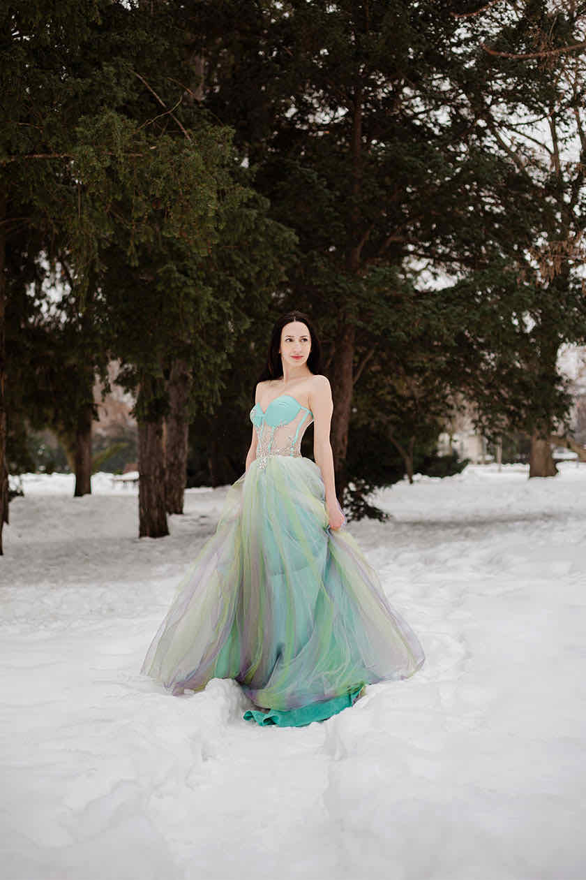 Woman in a pastel green and lilac tulle gown standing in snow, framed by dark pine trees